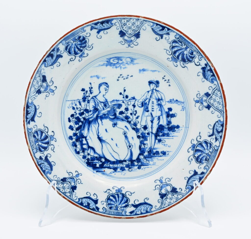 A Dutch Delft blue and white plate, painted with a pastoral scene depicting a gentleman playing a flute before a seated lady reading a letter. The subject belongs to the repertoire of Rococo genre scenes that became popular in Delft during the third quarter of the eighteenth century, reflecting contemporary taste for pastoral imagery derived from French prints and paintings. The broad rim is finished with a brown edge, a feature inspired by Chinese export porcelain. Brown-edged Delftware became particularly fashionable and favored in the mid-eighteenth century as Delft potteries responded to the continued popularity of Chinese porcelain among European consumers, adapting both decorative motifs and colour schemes to the Dutch tin-glazed tradition. The reverse bears the painted GVS mark of Geertruy Verstelle, proprietor of Het Oude Moriaenshooft between 1761 and 1773. Geertruy Verstelle occupies a distinctive place in the history of Delftware as one of the relatively few women to own and manage a Delft pottery in the eighteenth century. Following the death of her husband, Jacobus Halder, she assumed ownership of Het Oude Moriaenshooft in 1761, continuing the factory during a period of increasing competition from English creamware and imported porcelain. Like several widows active in the Delft ceramics trade, Verstelle directed both the commercial and artistic operations of the pottery. What distinguishes Verstelle from many of her contemporaries is that she marked her production with her own initials, GVS (Geertruy Verstelle), rather than continuing to use her late husband's mark or identifying herself simply as a widow (weduwe). This deliberate choice visibly asserted her own identity as proprietor and is an uncommon and significant feature in eighteenth-century Delftware. Pieces bearing her initials are therefore not only attributable to a well-defined period of production but also represent the independent leadership of one of the few documented female factory owners in Delft. Condition: Very good condition: Only slight wear consistent with age. Literature: C.H. de Jonge, Delfts Aardewerk (1965); J.D. van Dam, Delftse Porceleyne (2004)