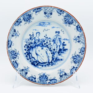 A Dutch Delft blue and white plate, painted with a pastoral scene depicting a gentleman playing a flute before a seated lady reading a letter. The subject belongs to the repertoire of Rococo genre scenes that became popular in Delft during the third quarter of the eighteenth century, reflecting contemporary taste for pastoral imagery derived from French prints and paintings. The broad rim is finished with a brown edge, a feature inspired by Chinese export porcelain. Brown-edged Delftware became particularly fashionable and favored in the mid-eighteenth century as Delft potteries responded to the continued popularity of Chinese porcelain among European consumers, adapting both decorative motifs and colour schemes to the Dutch tin-glazed tradition. The reverse bears the painted GVS mark of Geertruy Verstelle, proprietor of Het Oude Moriaenshooft between 1761 and 1773. Geertruy Verstelle occupies a distinctive place in the history of Delftware as one of the relatively few women to own and manage a Delft pottery in the eighteenth century. Following the death of her husband, Jacobus Halder, she assumed ownership of Het Oude Moriaenshooft in 1761, continuing the factory during a period of increasing competition from English creamware and imported porcelain. Like several widows active in the Delft ceramics trade, Verstelle directed both the commercial and artistic operations of the pottery. What distinguishes Verstelle from many of her contemporaries is that she marked her production with her own initials, GVS (Geertruy Verstelle), rather than continuing to use her late husband's mark or identifying herself simply as a widow (weduwe). This deliberate choice visibly asserted her own identity as proprietor and is an uncommon and significant feature in eighteenth-century Delftware. Pieces bearing her initials are therefore not only attributable to a well-defined period of production but also represent the independent leadership of one of the few documented female factory owners in Delft. Condition: Very good condition: Only slight wear consistent with age. Literature: C.H. de Jonge, Delfts Aardewerk (1965); J.D. van Dam, Delftse Porceleyne (2004)