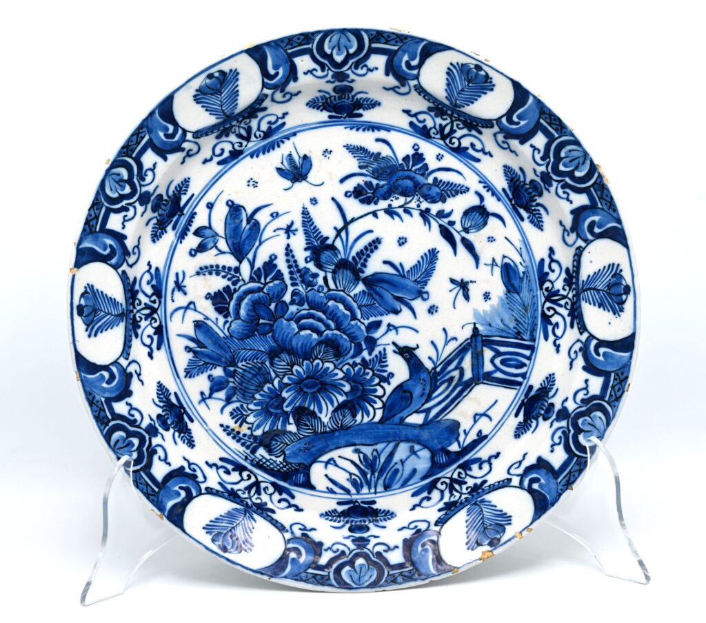 A Dutch Delft blue and white charger with chinoiserie decor of birds and insects, dating to circa 1720-1740. The central field is painted in cobalt blue with a bird beside flowering plants and a garden fence. Butterflies and other insects are distributed around the floral composition. The border is arranged in alternating sections of oval reserves containing stylised flowering sprigs, foliate motifs, diaperwork and smaller floral ornaments. The decoration reflects the continued influence of Chinese Kangxi porcelain on Delftware during the early 18th century. Chinese motifs such as birds, insects, flowering plants and garden architecture were combined and adapted by Delft painters to suit the broad surface of a large earthenware dish. The dense central composition and systematically divided border are characteristic of Delft chinoiserie decoration from the first half of the 18th century. The reverse is unmarked. Condition In good antique condition, with glaze wear, fritting and several small rim chips consistent with age. Size 35 cm Diameter