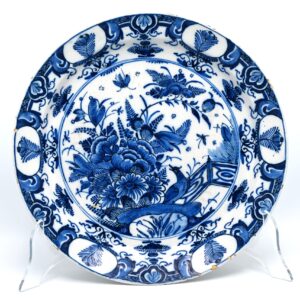 A Dutch Delft blue and white charger with chinoiserie decor of birds and insects, dating to circa 1720-1740. The central field is painted in cobalt blue with a bird beside flowering plants and a garden fence. Butterflies and other insects are distributed around the floral composition. The border is arranged in alternating sections of oval reserves containing stylised flowering sprigs, foliate motifs, diaperwork and smaller floral ornaments. The decoration reflects the continued influence of Chinese Kangxi porcelain on Delftware during the early 18th century. Chinese motifs such as birds, insects, flowering plants and garden architecture were combined and adapted by Delft painters to suit the broad surface of a large earthenware dish. The dense central composition and systematically divided border are characteristic of Delft chinoiserie decoration from the first half of the 18th century. The reverse is unmarked. Condition In good antique condition, with glaze wear, fritting and several small rim chips consistent with age. Size 35 cm Diameter