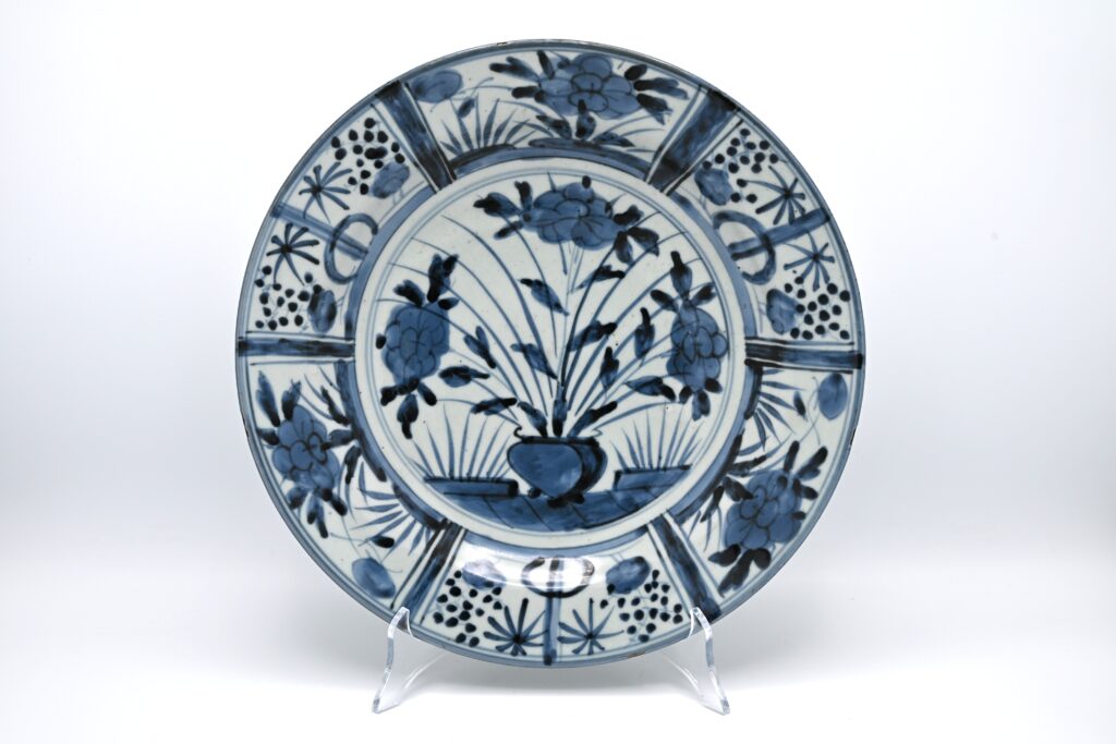 Antique 17th Century Japanese Arita Blue and White Kraak-Style Porcelain Charger, circa 1680 Product description Blue and White Arita Porcelain Charger in the Kraak Style Japan, Edo period, circa 1680 A Japanese Arita porcelain charger painted in underglaze cobalt blue. The central medallion depicts a flower basket on a fenced terrace, filled with flowering peonies and tall leafy stems. Two smaller plants flank the main arrangement. The broad rim is divided into six wide panels containing stylised bamboo, peony and prunus, separated by narrow radiating panels decorated with scrollwork in darker blue. The strong radial composition and alternating border panels were derived from Chinese Kraak porcelain produced for export during the late Ming dynasty. Following the disruption of Chinese porcelain exports in the mid-seventeenth century, Dutch merchants increasingly turned to the Japanese kilns at Arita. Japanese potters adapted familiar Chinese Kraak and Transitional designs for the European market, retaining their recognisable compositions while translating them into a distinctly Japanese style of painting. Large blue and white Arita dishes were exported through the Dutch East India Company during the second half of the seventeenth century. Comparable border designs also occur on Japanese porcelain bearing the VOC monogram and on a dated armorial dish from around 1667, supporting a date in the period 1660–1690 for this group. With its vigorous cobalt decoration, flower basket motif and Kraak-style panelled border, this antique Japanese porcelain charger is a characteristic example of early Arita export porcelain made for the Dutch market. Dating: circa 1680, within the documented 1660–1690 production period.