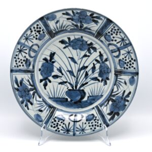 Antique 17th Century Japanese Arita Blue and White Kraak-Style Porcelain Charger, circa 1680 Product description Blue and White Arita Porcelain Charger in the Kraak Style Japan, Edo period, circa 1680 A Japanese Arita porcelain charger painted in underglaze cobalt blue. The central medallion depicts a flower basket on a fenced terrace, filled with flowering peonies and tall leafy stems. Two smaller plants flank the main arrangement. The broad rim is divided into six wide panels containing stylised bamboo, peony and prunus, separated by narrow radiating panels decorated with scrollwork in darker blue. The strong radial composition and alternating border panels were derived from Chinese Kraak porcelain produced for export during the late Ming dynasty. Following the disruption of Chinese porcelain exports in the mid-seventeenth century, Dutch merchants increasingly turned to the Japanese kilns at Arita. Japanese potters adapted familiar Chinese Kraak and Transitional designs for the European market, retaining their recognisable compositions while translating them into a distinctly Japanese style of painting. Large blue and white Arita dishes were exported through the Dutch East India Company during the second half of the seventeenth century. Comparable border designs also occur on Japanese porcelain bearing the VOC monogram and on a dated armorial dish from around 1667, supporting a date in the period 1660–1690 for this group. With its vigorous cobalt decoration, flower basket motif and Kraak-style panelled border, this antique Japanese porcelain charger is a characteristic example of early Arita export porcelain made for the Dutch market. Dating: circa 1680, within the documented 1660–1690 production period.