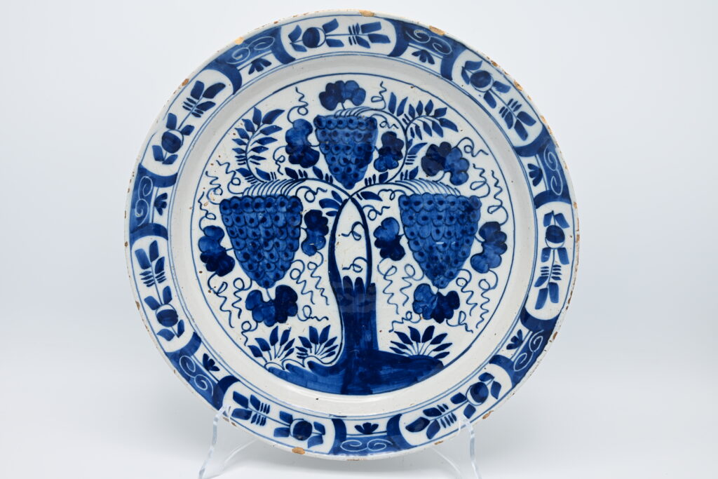 18th-Century Dutch Delft Blue and White Dish with Grapevine An 18th-century Dutch Delftware dish painted in cobalt blue with three prominent bunches of grapes growing from a stylised vine. Smaller grapes, leaves and curling tendrils fill the central field, while the border is divided into panels of stylised floral motifs and scrolling ornament. The dish retains an old collection label numbered 546. Tin-glazed earthenware. Unmarked. Delft, the Netherlands, 18th century. Provenance: Formerly in the collection of Henri Blay (1861–1920). Condition: Small chips, glaze losses and scratches around the rim, consistent with age. Diameter: 30.5 cm.