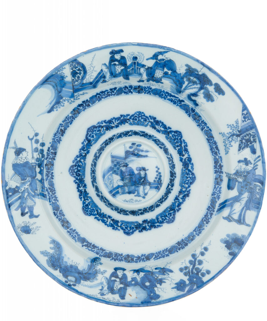 A large Dutch Delftware rosewater dish, painted in cobalt blue on an opaque white tin glaze. The moulded form consists of a shallow central well with a domed centre, surrounded by raised concentric bands and an exaggerated broad rim. The domed central medallion depicts two Chinese figures seated on a garden terrace, with a pavilion and mountainous landscape in the distance. It is surrounded by dense bands of flowering plants, foliate scrollwork and lappet-shaped ornament. The broad rim is painted with four figural scenes showing Chinese dignitaries, scholars and attendants among pierced rocks, pine trees, shrubs and garden fences. The decoration is derived from Chinese Transitional porcelain of the seventeenth century. Rather than following a single porcelain prototype, the Delft painter combined various Asian figures and landscape motifs within a distinctly European form. Rosewater dishes were made to contain rosewater or other scented liquids. These could be used to refresh or cleanse the hands, or to perfume the surrounding air. Their large dimensions, elaborate decoration and association with refined dining customs also made them suitable for prominent display. The reverse is decorated in blue with a repeated pattern of stylised circular motifs separated by vertical lines. Unmarked. The form belongs to a group of large Delft rosewater dishes dating from the final quarter of the seventeenth century. Closely related examples from circa 1675–1680 have been attributed to Samuel van Eenhoorn, owner of De Grieksche A from 1678 until 1686. Without a factory mark, however, an attribution of the present dish remains uncertain. Delft, circa 1680.