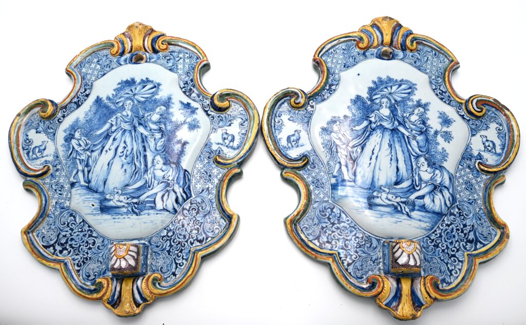 A pair of mid-18th-century Dutch Delftware wall plaques with blue-painted biblical scenes and polychrome moulded borders. Each plaque has a suspension hole at the top and a projecting socket below, made to receive a candle arm. The pair was designed as wall sconces, combining candlelight with a narrative image in tin-glazed earthenware. The plaques depict the Finding of Moses (Exodus 2:5–6). Pharaoh’s daughter stands beneath a parasol beside the river as an attendant bends towards the infant in his basket. The central scenes are framed by scrollwork and floral ornament picked out in yellow, green and manganese, with small panels depicting sheep on either side. Both plaques follow the same composition, with slight differences in the painted details. The scene derives from one of six Old Testament subjects designed by Jacopo Amigoni (1682–1752) and circulated as engravings published by Joseph Wagner in Venice. The other subjects include David and Abigail, Rebecca at the Well, Moses Striking a Shepherd, Abraham Kneeling before the Three Angels and Jacob’s Ladder. The National Gallery of Art, Washington, holds a complete set (inv. nos. 1984.84.1–1984.84.6), including The Finding of Moses, inv. no. 1984.84.1. www.nga.gov Similar examples: A Delftware plaque with the same subject is in the Rijksmuseum, Amsterdam, inv. no. BK-1958-34-A. Another is in the Cleveland Museum of Art, inv. no. 2024.12. www.rijksmuseum.nl Literature relating to the Rijksmuseum example: Henry Havard, Catalogue chronologique et raisonné des faïences de Delft composant la collection de John F. Loudon (1877), no. 418, p. 71; T. G. Kootte, ed., De bijbel in huis (1991), no. 146, p. 100; Jan Daniël van Dam, Delffse Porceleyne: Delfts aardewerk 1620–1850 (2004), pp. 138–139.