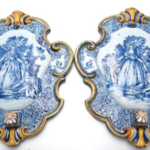 A pair of mid-18th-century Dutch Delftware wall plaques with blue-painted biblical scenes and polychrome moulded borders. Each plaque has a suspension hole at the top and a projecting socket below, made to receive a candle arm. The pair was designed as wall sconces, combining candlelight with a narrative image in tin-glazed earthenware. The plaques depict the Finding of Moses (Exodus 2:5–6). Pharaoh’s daughter stands beneath a parasol beside the river as an attendant bends towards the infant in his basket. The central scenes are framed by scrollwork and floral ornament picked out in yellow, green and manganese, with small panels depicting sheep on either side. Both plaques follow the same composition, with slight differences in the painted details. The scene derives from one of six Old Testament subjects designed by Jacopo Amigoni (1682–1752) and circulated as engravings published by Joseph Wagner in Venice. The other subjects include David and Abigail, Rebecca at the Well, Moses Striking a Shepherd, Abraham Kneeling before the Three Angels and Jacob’s Ladder. The National Gallery of Art, Washington, holds a complete set (inv. nos. 1984.84.1–1984.84.6), including The Finding of Moses, inv. no. 1984.84.1. www.nga.gov Similar examples: A Delftware plaque with the same subject is in the Rijksmuseum, Amsterdam, inv. no. BK-1958-34-A. Another is in the Cleveland Museum of Art, inv. no. 2024.12. www.rijksmuseum.nl Literature relating to the Rijksmuseum example: Henry Havard, Catalogue chronologique et raisonné des faïences de Delft composant la collection de John F. Loudon (1877), no. 418, p. 71; T. G. Kootte, ed., De bijbel in huis (1991), no. 146, p. 100; Jan Daniël van Dam, Delffse Porceleyne: Delfts aardewerk 1620–1850 (2004), pp. 138–139.